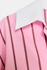 Camisa oversize de popelina rosa con rayas verticales marrones, cuello solapa blanco y escote en pico. Mangas largas con puños anchos blancos a contraste y cierre frontal con botones.