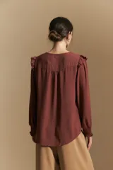 Camisa negra de manga larga con escote en V y cierre frontal de botones. Presenta detalles de volados en los hombros, frunces en la espalda y puños elastizados que le otorgan una silueta relajada y fluida.