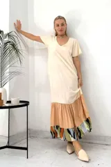 Vestido largo de manga corta con cuello en V, de color crema liso en la parte superior. La falda es de corte amplio con un volante en color melocotón liso, y un volante inferior con estampado abstracto en tonos verde oliva, negro, naranja y azul claro.