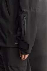 Cazadora deportiva de esquí en color negro, confeccionada en tejido técnico impermeable de tres capas con costuras y cremalleras termoselladas. Cuenta con capucha ajustable, ventilación en axilas, puños con ajuste de tira adherente y puño interior elástico con abertura para el pulgar. Incluye sistema de rescate RECCO y faldón interior para protección contra el frío.