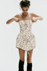 Vestido corto strapless de corte corsetero, con estampado floral en tonos rosados y verdes sobre fondo blanco. Presenta detalles de tiras cruzadas ajustables en el delantero con lazada y varillas interiores.