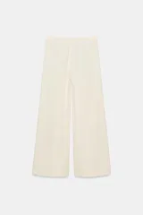 Pantalón wide leg de tiro medio confeccionado en hilatura de algodón, color beige, con trabillas en la cintura, bolsillos laterales ocultos y detalle de pliegues en el delantero. Cierre frontal con cremallera, botón y ganchos metálicos.