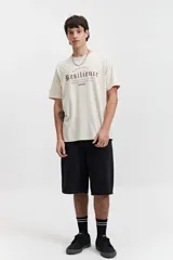 Remera de manga corta color beige, con escote redondo y fit regular. Presenta un estampado frontal en color marrón con la palabra "Resilience" en tipografía gótica y el texto "Original The World Has Changed Established Since 1976 // NLD Mistral".