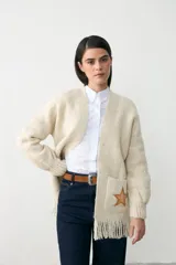 Chaqueta de punto color crema, con diseño abierto, mangas largas y flecos en el ruedo. Presenta un aplique bordado en forma de estrella en uno de los bolsillos frontales.