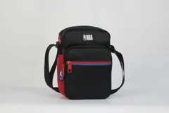 Morral pequeño tipo bandolera, color negro, con detalles en rojo y azul en el frente. Presenta el logo de la NBA en la parte superior y un bolsillo frontal con cierre. Incluye correa regulable.
