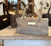 Cartera tipo tote bag de formato rectangular y alargado, confeccionada en gamuza color marrón oscuro. Presenta doble asa de hombro, solapa frontal con cierre metálico giratorio en tono dorado y costuras visibles en los bordes.