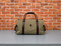 Bolso tipo duffel de lona color verde oliva con detalles en cuero marrón oscuro. Cuenta con asas superiores reforzadas, correas verticales de cuero y un pequeño parche circular con logo en el centro.