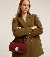 Cartera tipo shoulder bag de tamaño mediano, confeccionada en poliuretano color marrón oscuro. Presenta una solapa frontal con un detalle metálico dorado de forma orgánica y asa ajustable.