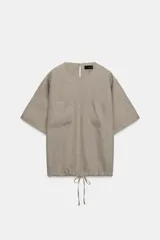 Top de lino de color beige, con cuello redondo, manga corta, bolsillos frontales aplicados y bajo ajustable con cordones. Cuenta con abertura en la espalda y cierre de botón.