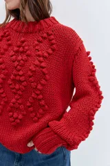 Sweater de punto grueso color rojo, con cuello alto y diseño de relieves tipo pompones en el frente y las mangas. Presenta puños y ruedo acanalados.