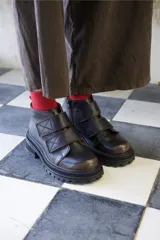 Borcego de cuero color marrón oscuro con acabado desgastado en la puntera. Presenta un diseño de caña corta con cierre lateral y dos tiras ajustables con velcro en el empeine. Cuenta con una suela de goma robusta tipo tractor.