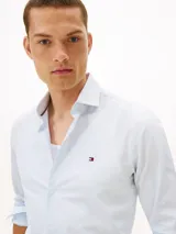 Camisa de Tommy Hilfiger confeccionada en popelín de puro algodón regenerativo con diseño de rayas bengal en tonos celeste y blanco. Cuenta con cuello camisero, cierre frontal de botones y el logo de la marca bordado en el pecho.