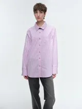 Camisa de corte oversize confeccionada en popelina de algodón, con diseño de rayas verticales en tonos rosa y blanco. Presenta cuello clásico, cierre frontal con botones, mangas largas con puños abotonados y un bolsillo aplicado en el pecho.