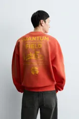 Sudadera de corte relajado, color naranja con efecto tie-dye/deslavado, cuello redondo y manga larga. Presenta un estampado frontal con texto y un trébol de cuatro hojas en amarillo.