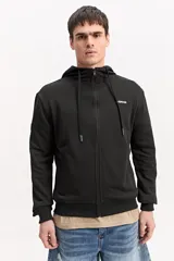 Campera de algodón gris melange con capucha, cierre frontal completo y bolsillos laterales. Cuenta con un parche rectangular negro con inscripciones en la manga y cordones ajustables para la capucha.