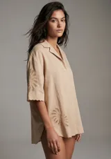 Camisa de manga corta color beige, con cuello camisero y botones frontales. Presenta bordados calados con diseño de hojas o abanicos en las mangas y en la parte inferior del frente. El ruedo es festoneado.