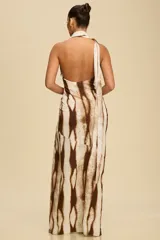 Vestido largo de satén con estampado abstracto en tonos marrones y beige. Presenta un diseño asimétrico con cuello halter tipo corbatín, espalda descubierta y un tajo profundo en la pierna con detalle drapeado en la cadera.