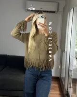 Cárdigan de tejido de punto color crema, con cuello redondo, mangas largas y cierre frontal con botones metálicos. Presenta detalles de flecos en los puños y en el ruedo.
