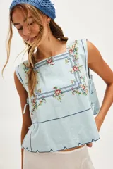 Top celeste sin mangas de algodón con escote cuadrado, bordado floral multicolor y líneas decorativas oscuras. Presenta un ruedo festoneado y laterales abiertos con tiras para ajustar.