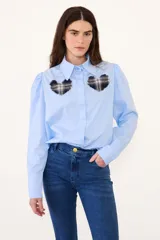 Camisa de algodón color celeste, con cuello clásico, mangas largas con hombros ligeramente abullonados y cierre frontal oculto. Presenta dos apliques en forma de corazón con estampado de cuadros escoceses y bordes desflecados en el pecho.