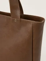 Bolso tipo tote de cuero color marrón, con diseño minimalista, doble asa de mano y cierre superior. Presenta el logo de la marca grabado en un parche central y detalles de remaches metálicos.