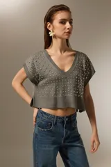 Chaleco de punto gris con cuello en V, diseño sin mangas y corte cropped. Presenta apliques de tachas metálicas plateadas distribuidas por toda la parte frontal y borde acanalado en el ruedo.