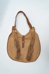 Bolso tipo hobo de gamuza color camel, con flecos largos laterales, correa de hombro de cuero sintético y cierre central con solapa y botón a presión.