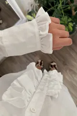 Camisa blanca de algodón con cuello clásico, cierre frontal con botones, mangas largas con puños abotonados y volados en el frente y los puños.