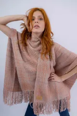 Ruana artesanal tejida en telar manual, con textura rústica y terminación en flecos. Presenta un diseño liviano en tonos rosados y naranjas, con etiqueta de marca en el frente.