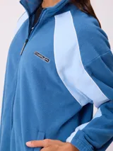 Campera de polar marca Rusty, color azul con paneles laterales en celeste claro. Presenta cierre frontal completo, cuello alto y logo de la marca bordado en el pecho.