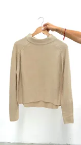 Sweater de punto en color beige, con cuello redondo ligeramente elevado y mangas largas con costura raglán. Presenta un diseño de corte recto con un ruedo asimétrico, siendo más corto en la parte frontal.