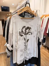 Remera de línea italiana con calce holgado, cuello redondo y mangas con puños vueltos que revelan un detalle a rayas. Presenta un estampado floral central con detalles dorados y texto, además de tiras ajustables en el ruedo inferior.