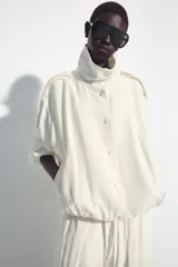 Chaqueta oversize negra confeccionada en hilatura con mezcla de modal, de corte fluido. Cuenta con cuello subido, manga larga con puños elásticos, bolsillos de vivo en el delantero y trabillas en los hombros. El bajo es abullonado con elástico y el cierre frontal es con botones a presión.