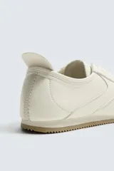 Zapatilla de estilo retro con cordones, confeccionada en una combinación de piezas de cuero sintético negro y detalles en blanco, con suela dentada de goma en tono caramelo.