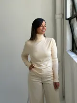 Polera de punto color crudo, con cuello alto y mangas largas que presentan puños acanalados con detalle de botones dorados.