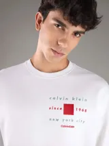 Buzo Calvin Klein blanco de manga larga, confeccionado en algodón regenerado y poliéster reprocesado con interior de felpa suave. Presenta cuello redondo, puños y dobladillo acanalados, y un estampado frontal con texto y un cuadrado rojo.