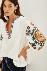 Camisa blanca de doble gasa de algodón, corte amplio y mangas largas abullonadas. Presenta bordados a mano en mangas y escote con motivos florales en verde, amarillo y violeta.