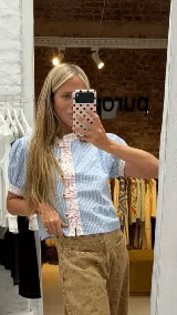 Blusa celeste a rayas blancas, de manga corta abullonada. Cuenta con cuello mao y tapeta de botones con detalles de estampado floreado en tono rosado, presentes también en el cuello y los puños.