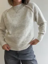 Sweater de punto de corte holgado en color gris melange, con cuello redondo y puños acanalados que presentan un detalle de contraste en color blanco. Posee mangas tipo raglán y una textura suave y elástica.