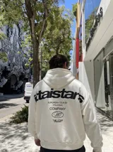Canguro blanco con capucha, con gran estampado en la espalda en color negro que incluye texto y logotipos de la marca.