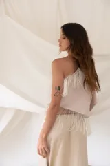 Top beige asimétrico de un solo hombro con mangas murciélago amplias, adornado con detalles de flecos delicados en el escote y en el ruedo inferior.