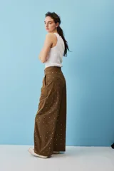 Pantalón marrón de corte ancho, tiro alto y diseño holgado, adornado con pequeños apliques metálicos distribuidos por toda la prenda.