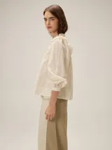 Camisa de color crema con bordados florales, cuello mao, cierre frontal con botones y mangas largas abullonadas con volados en los hombros.
