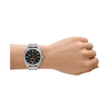 Reloj cronógrafo Fossil Machine con caja de 42 mm y malla de acero inoxidable plateado. Presenta una esfera negra con detalles en naranja, manecillas luminiscentes y movimiento de cuarzo.