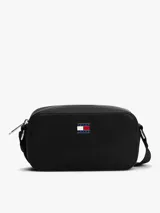 Cartera tipo bandolera de diseño compacto y minimalista, confeccionada en poliuretano con acabado liso. Presenta cierre de cremallera superior y una insignia metálica distintiva de Tommy Jeans en el centro del frente.