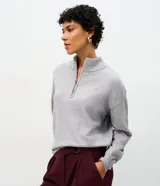 Suéter de punto color gris con cuello alto y medio cierre frontal. Presenta mangas largas con puños ajustados y una sutil aplicación de brillos en la parte frontal.