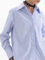 Camisa de poplin color blanco, con cuello clásico, cierre frontal de botones y mangas largas con puños. Presenta un diseño bordado de pequeñas llamas distribuidas por toda la prenda.