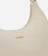 Cartera tipo baguette de diseño minimalista en color beige, confeccionada en poliuretano con acabado liso. Presenta cierre superior con cremallera, asa ajustable y detalles metálicos en tono dorado claro, incluyendo una placa con el logo de la marca en el frente.