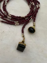 Collar largo de cuentas pequeñas en tono bordó, intercaladas con esferas doradas. Presenta dos dijes colgantes con piedras negras y detalles metálicos dorados. Su diseño permite usarlo largo o con doble vuelta.