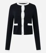 Cardigan corto de punto en color blanco con detalles en negro, cuello redondo, manga larga y cierre frontal de botones perlados con ribete negro. Cuenta con dos bolsillos delanteros de ojal con ribete negro y botón.